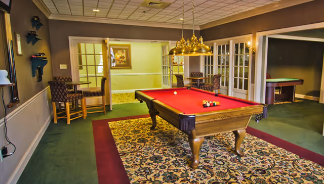A recreational room with a red felt pool table centered on a patterned carpet. The room has green carpet flooring with a maroon border around the rug. There are two sets of French doors leading to adjacent rooms, one with additional seating and another with a second game table. The walls are painted in neutral tones, and a brass light fixture with multiple lamps hangs above the pool table. Chairs and small tables are arranged near the doors.
