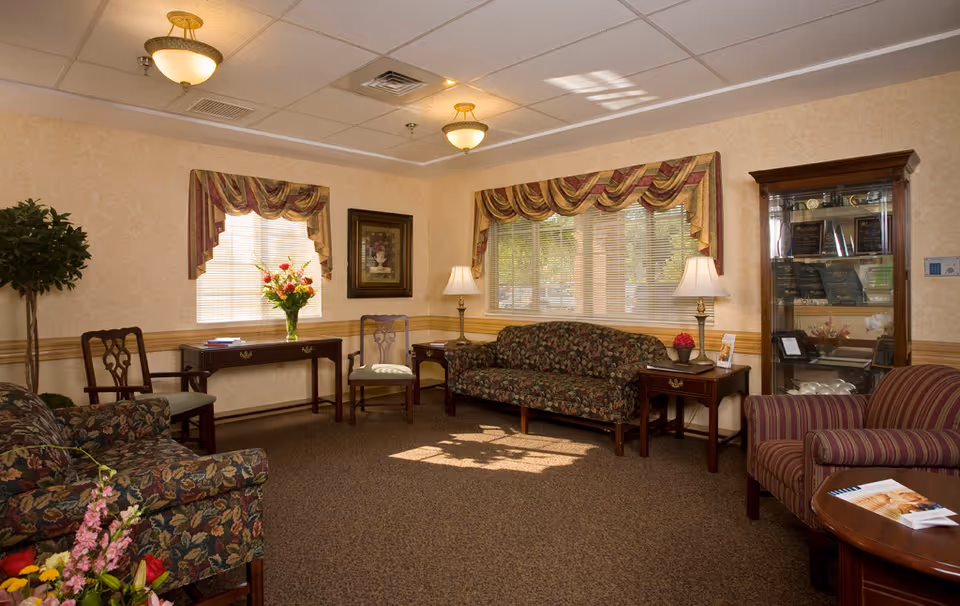 A cozy living room area with floral patterned sofas and chairs, a wooden coffee table with magazines, two side tables with lamps, a wooden display cabinet with awards and decorative items, and windows with striped valances letting in natural light.