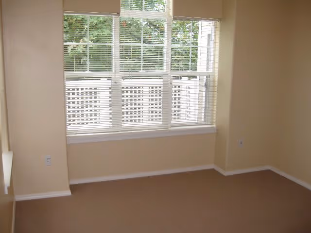 Empty room with beige walls and carpet, featuring a large window with white blinds partially open, allowing a view of greenery outside.