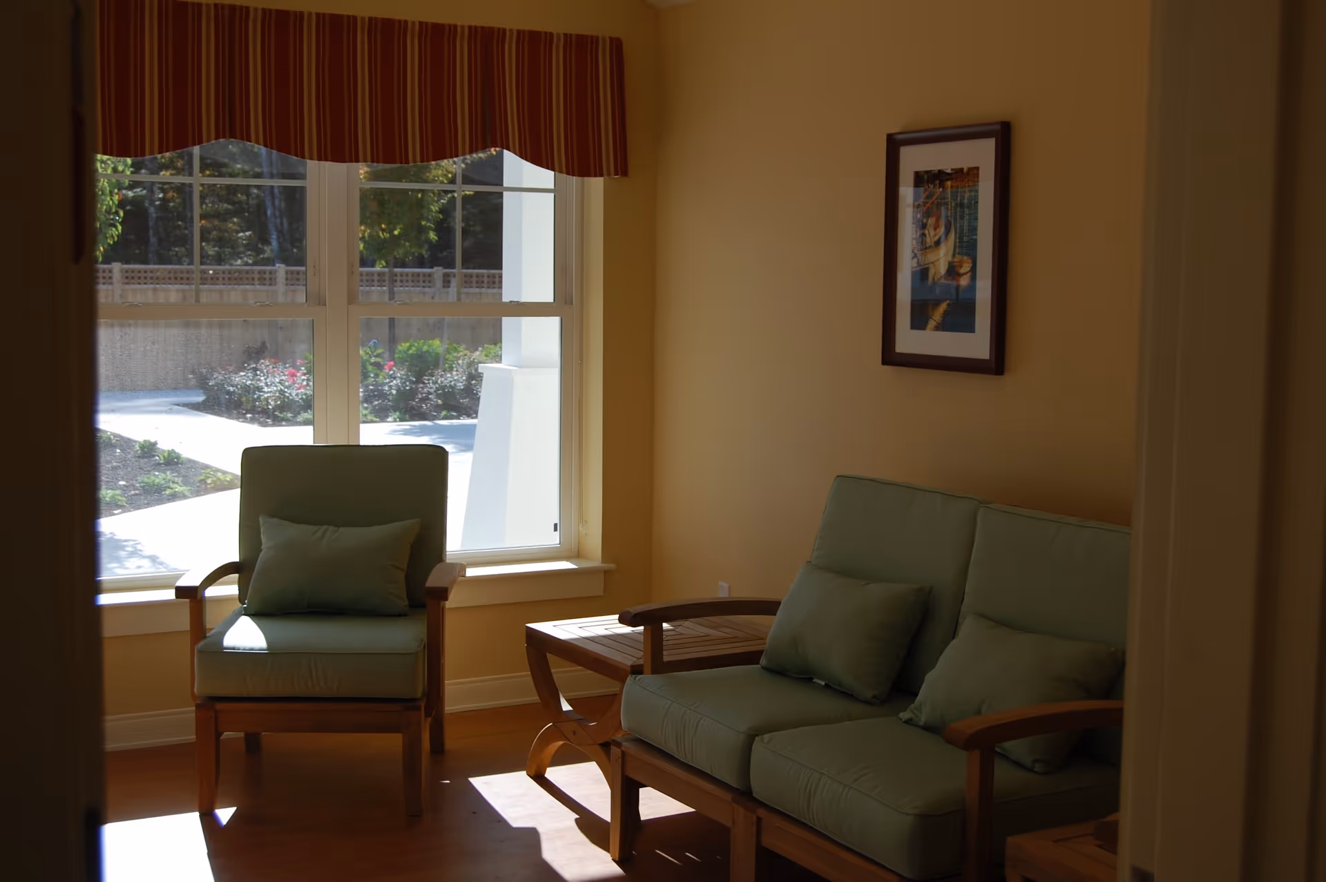 Sunlit seating area with green cushioned armchairs and a loveseat by a large window with a striped valance.