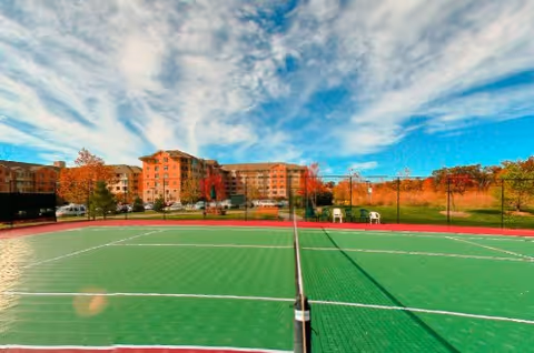 Outdoor tennis court with a net in the foreground and a large multi-story senior living community building in the background under a partly cloudy blue sky. Trees with autumn foliage surround the area.