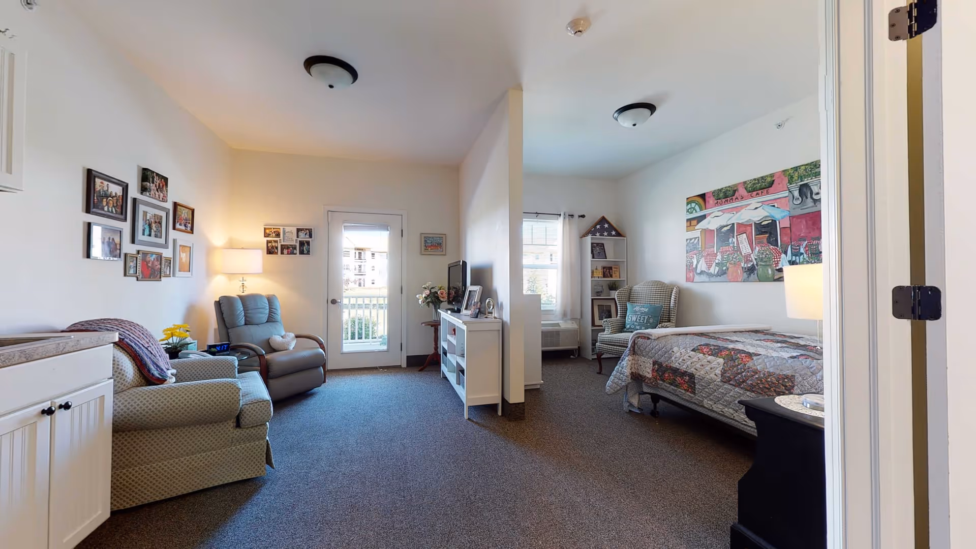 A cozy and well-lit assisted living room featuring a sitting area with two armchairs, a small table with a lamp, and family photos on the wall. The room is divided by a partial wall, with a bed, a cushioned chair, a bookshelf, and a colorful painting on the opposite side. A door with a window leads to an outside balcony or patio.
