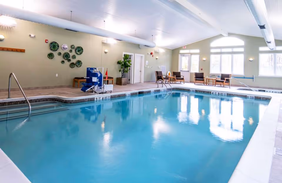 Indoor swimming pool area with clear blue water, metal handrails, pool lift chair, several chairs and tables along the walls, large windows letting in natural light, and decorative wall art.