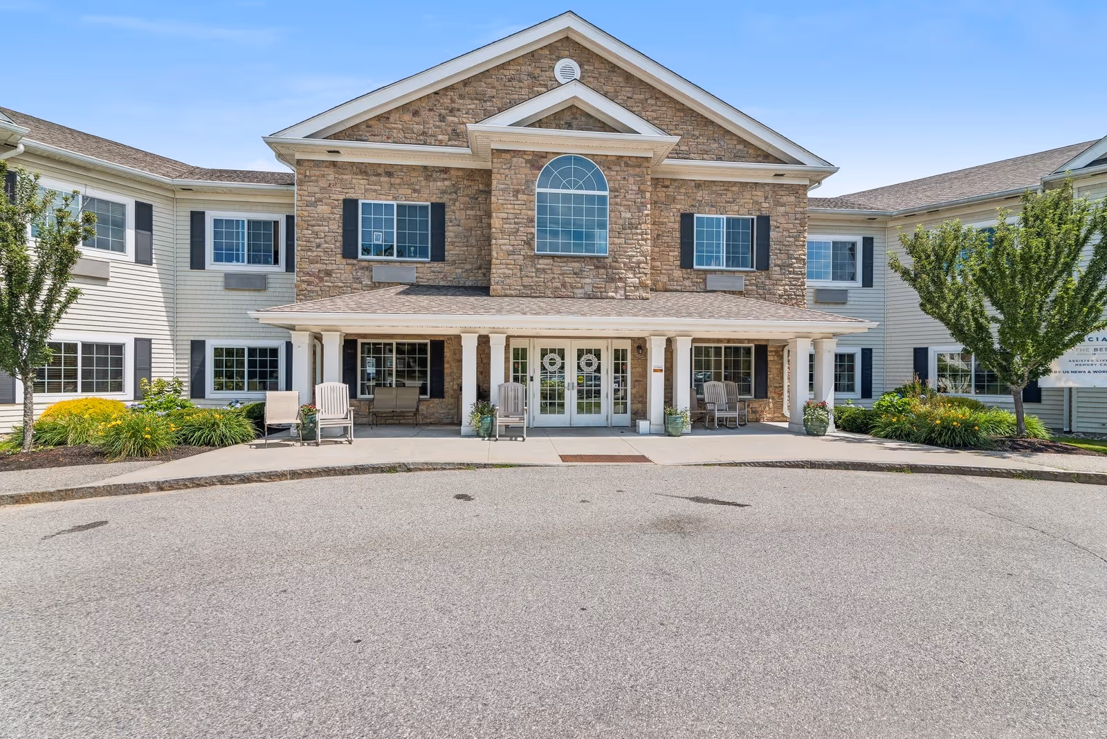 Front exterior view of The Enclave of Scarborough building with stone and siding facade, multiple windows with black shutters, a covered entrance with white columns, and outdoor chairs on the porch. There are landscaped bushes and trees on either side of the entrance under a clear blue sky.