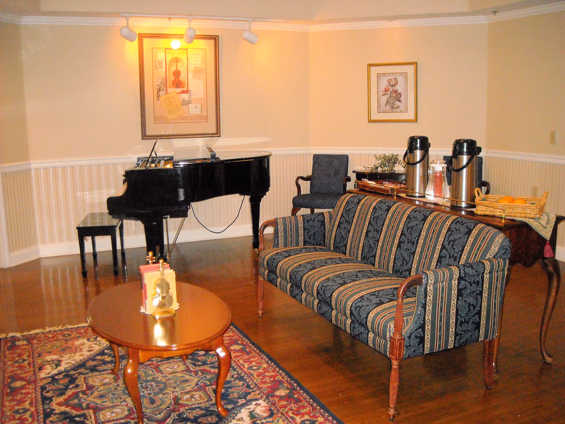 A cozy living room area with a patterned upholstered sofa, a wooden coffee table with decorative items, a grand piano with a matching bench, a side table with two large beverage dispensers and a basket of oranges, and framed artwork on the walls.