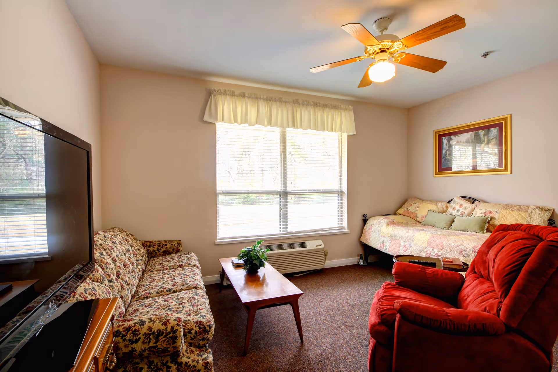 A cozy living room with a floral patterned sofa, a red recliner chair, a wooden coffee table with a small plant, a daybed with multiple cushions, a large window with blinds and a valance, a ceiling fan with a light, and a flat-screen TV on a wooden stand.