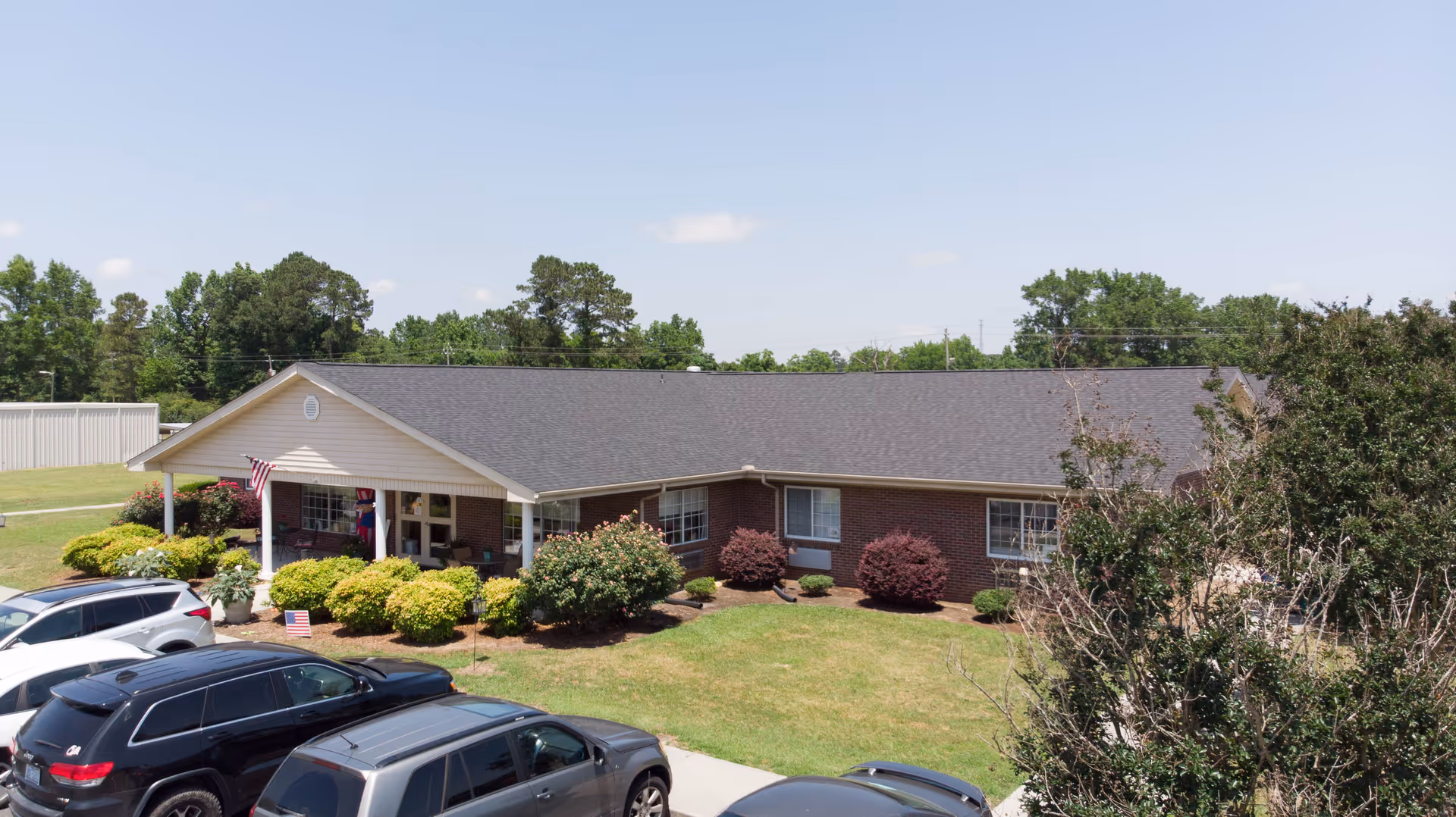 Exterior view of a single-story brick building with a dark shingled roof, surrounded by green bushes and trees. Several cars are parked in front of the building under a clear blue sky.