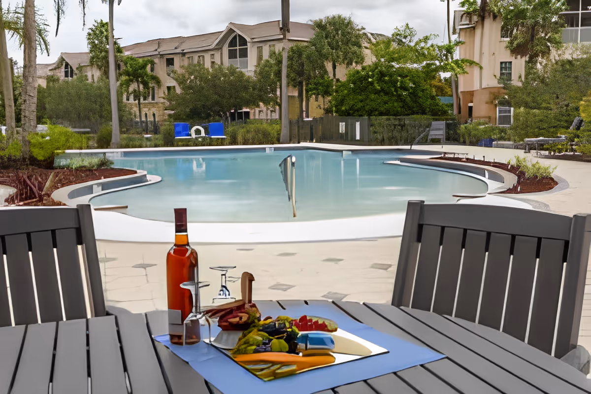 Outdoor pool area with a curved swimming pool surrounded by a paved deck. In the foreground, there is a wooden table with two chairs, a bottle of wine, two wine glasses, and a platter of assorted fruits and vegetables. Trees and buildings are visible in the background under a cloudy sky.