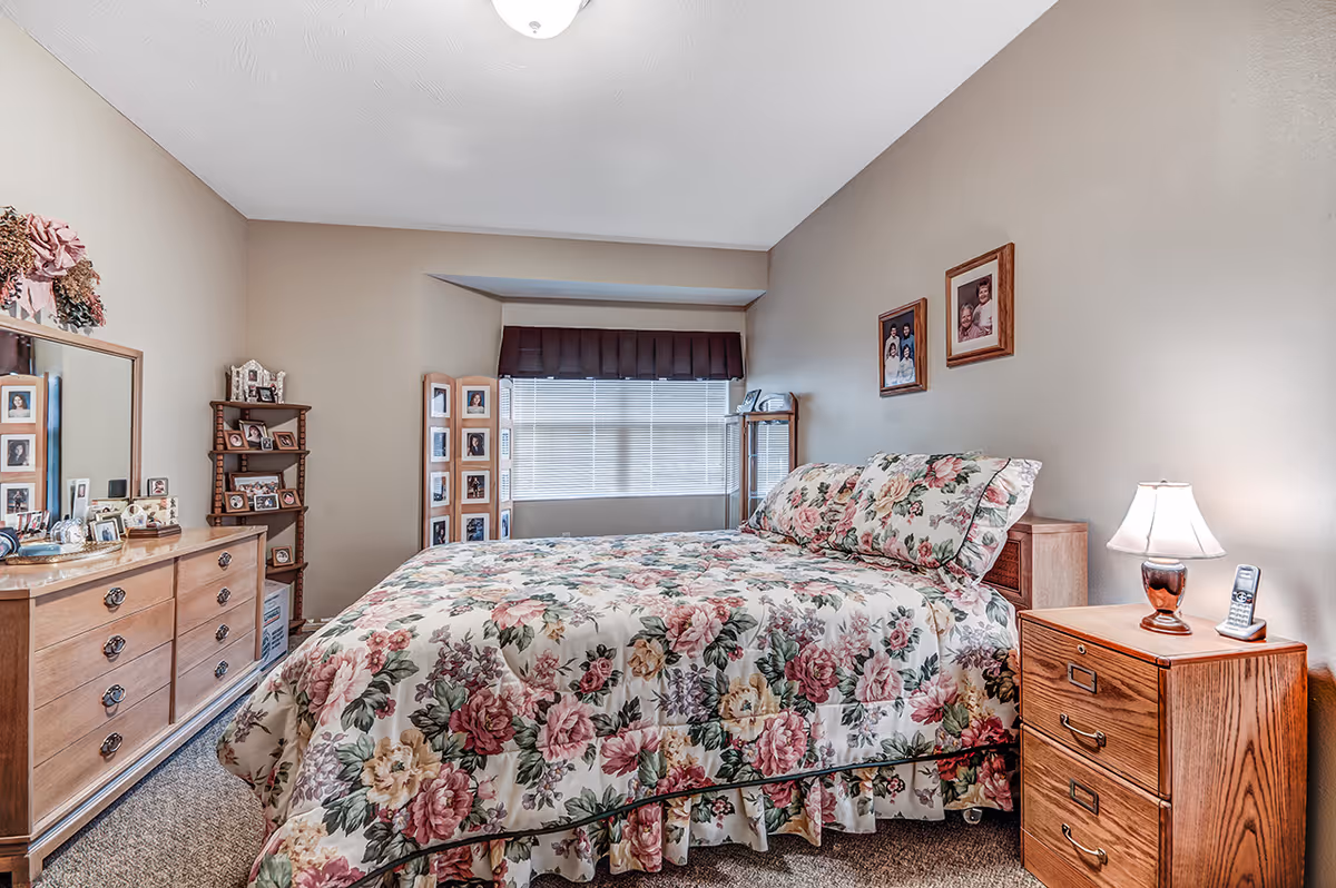 A bedroom with a bed covered in a floral bedspread and matching pillows. There is a wooden nightstand with a lamp and a cordless phone on it next to the bed. On the opposite side, there is a wooden dresser with a large mirror and various framed photos on top. A small wooden shelf with more framed photos is in the corner. The walls are beige, and there are framed pictures hanging above the bed. A window with blinds and a dark valance is at the far end of the room.