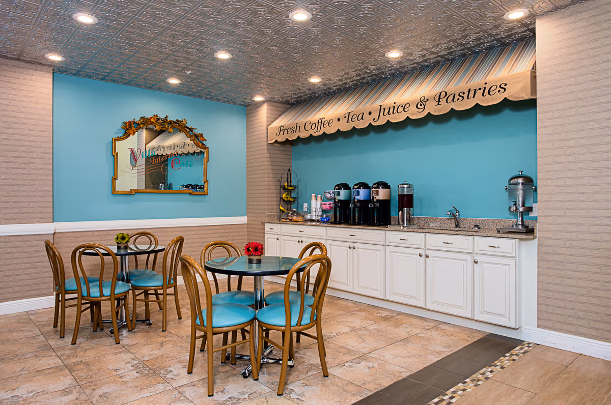 A cozy refreshment area with two round tables and eight wooden chairs with teal cushions. The back wall is painted teal and features a counter with multiple beverage dispensers and a sink. Above the counter is a striped awning with the words 'Fresh Coffee • Tea • Juice & Pastries'. A decorative mirror hangs on the adjacent wall, reflecting part of the room.