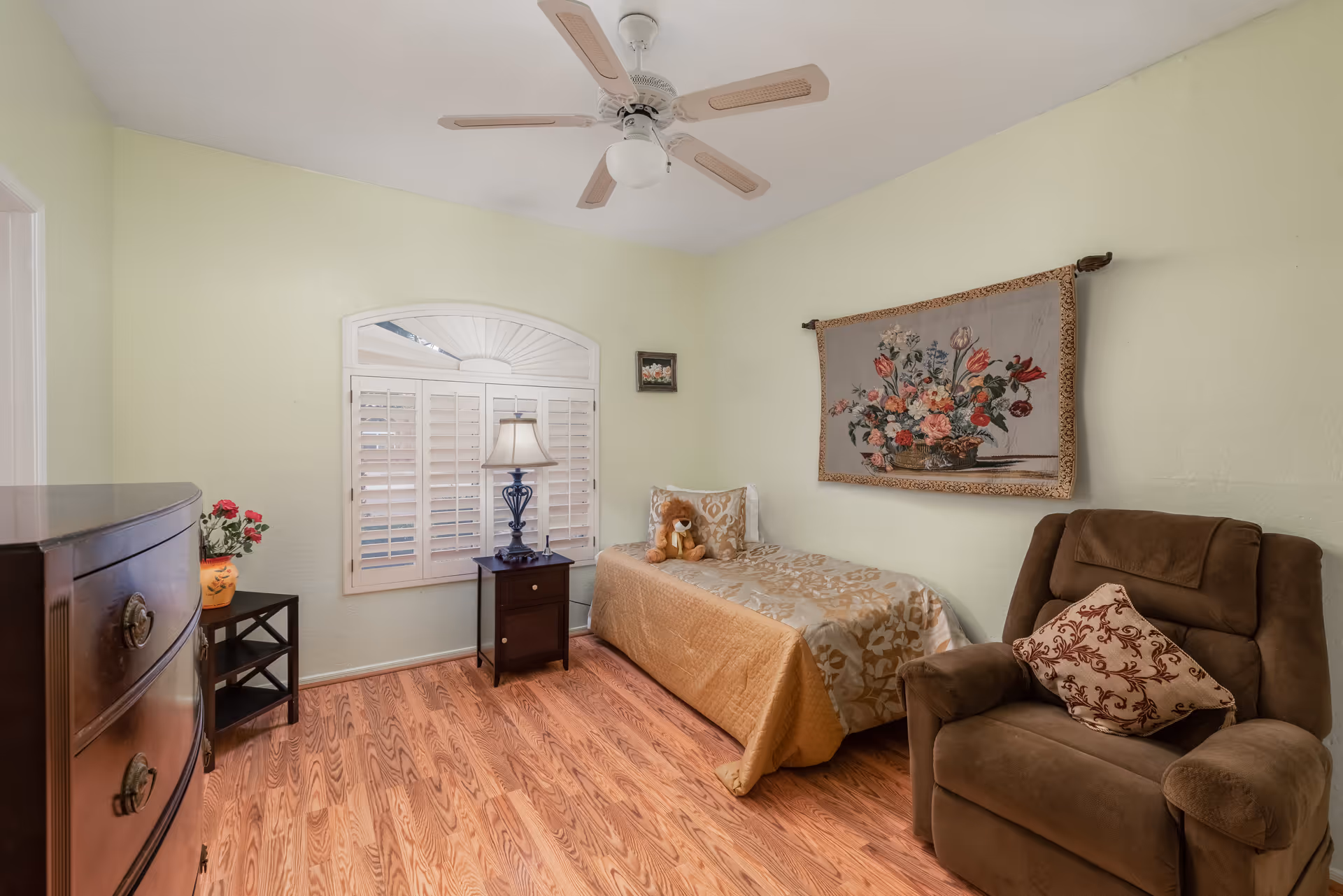 A small bedroom with light green walls and wood flooring. The room features a single bed with a gold and beige patterned bedspread, a teddy bear on the bed, a brown cushioned armchair with a decorative pillow, a dark wooden dresser, a small side table with a lamp, and a window with white plantation shutters. A floral tapestry hangs on the wall above the bed, and a ceiling fan with light is mounted on the ceiling.
