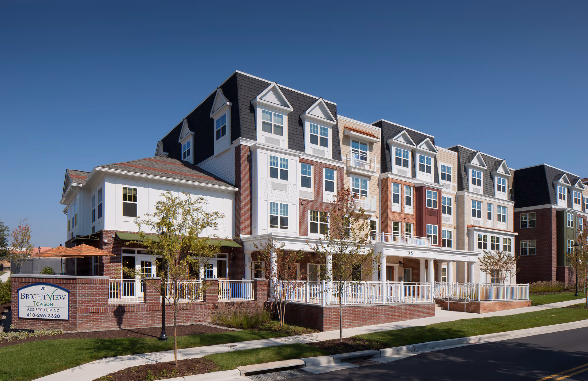 Front exterior of a multi-story senior living building with brick and siding, balconies, and a fenced entrance under a clear blue sky.