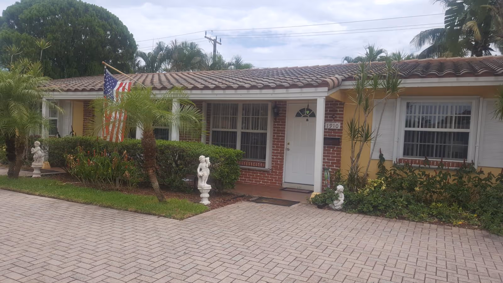 Front exterior view of a single-story residential building with a tiled roof, brick and yellow walls, white door, and windows with white shutters. The entrance is flanked by small palm trees, bushes, and decorative statues. An American flag is displayed near the entrance.