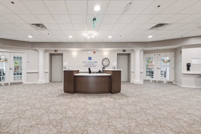 Reception area of Fox Hollow Senior Living Community featuring a central wooden reception desk, two elevators on either side, a wall clock, and glass doors leading to other rooms. The space has a patterned carpet, white walls, and ceiling lights.