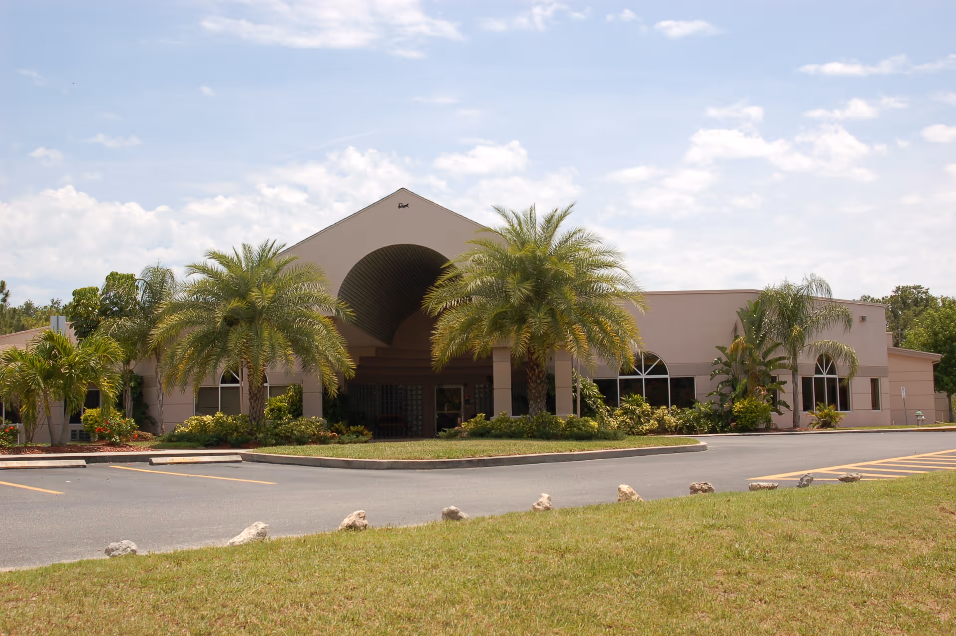 Exterior view of Surrey Place Healthcare & Rehabilitation building with a covered entrance, palm trees, and landscaped greenery under a partly cloudy sky.