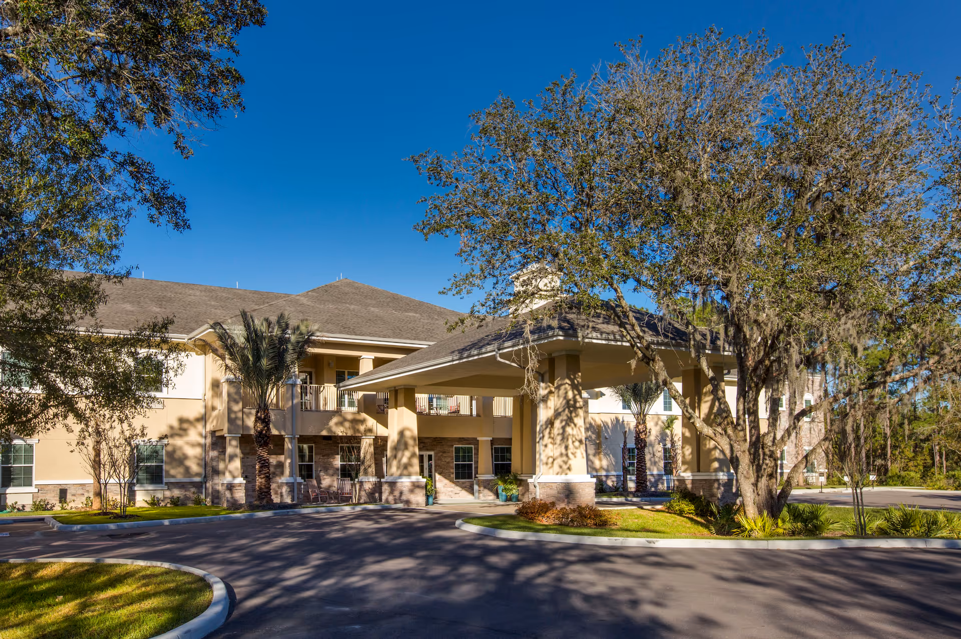 Exterior view of The Legacy at Highwoods Preserve senior living facility showing a two-story building with a covered entrance, surrounded by trees and landscaping under a clear blue sky.
