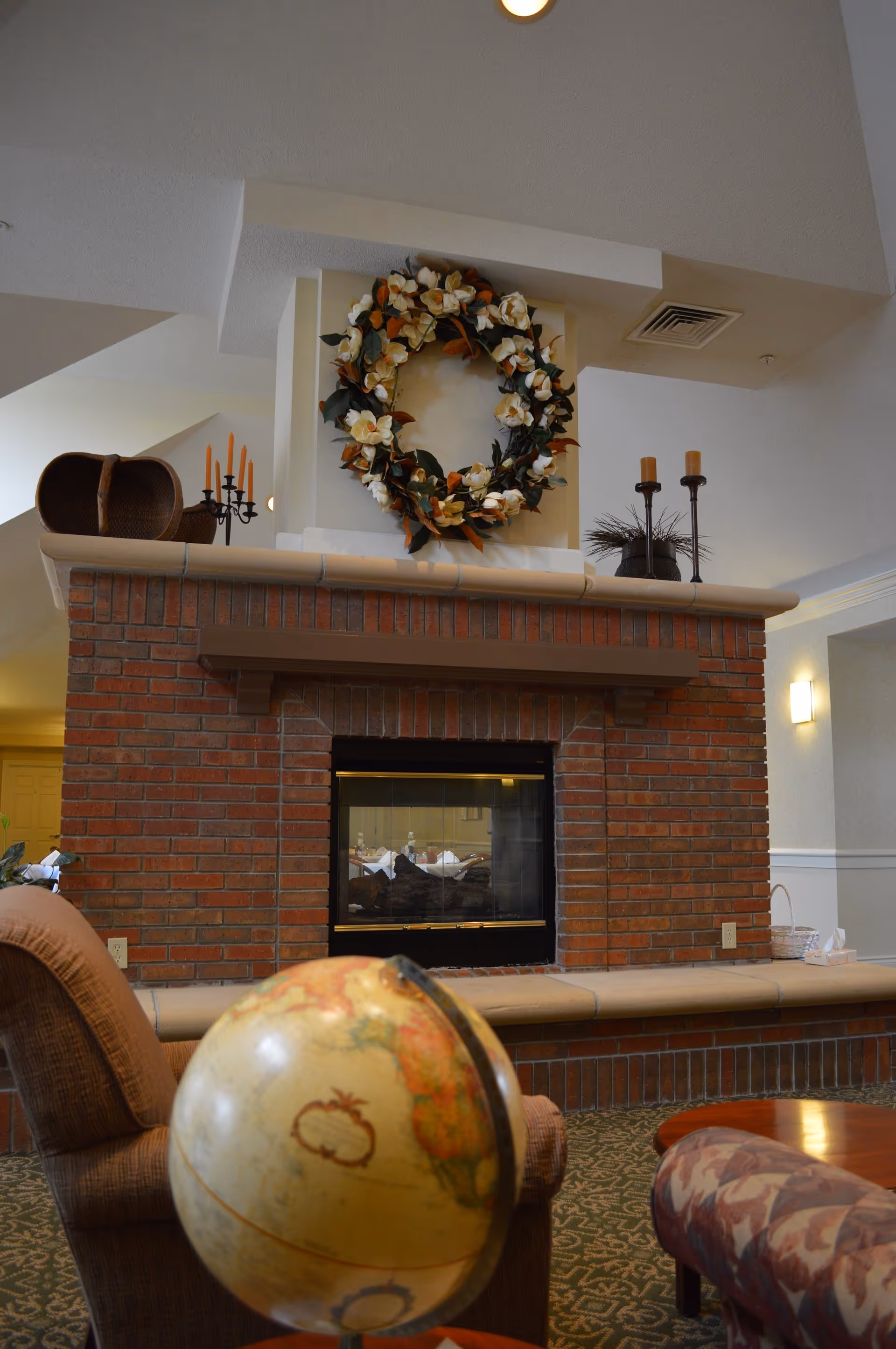 Cozy seating area with a brick double-sided fireplace topped by a decorative wreath and candlesticks, with a globe and chairs in the foreground.