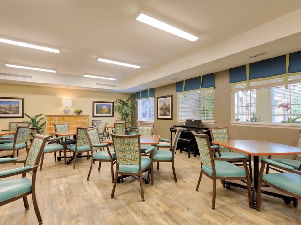 A bright and spacious dining room with multiple wooden tables and green cushioned chairs arranged neatly. The room has large windows with blue valances, framed pictures on the walls, a wooden sideboard with a lamp and plants, and a piano in the corner. The floor is light wood, and the ceiling has recessed lighting.