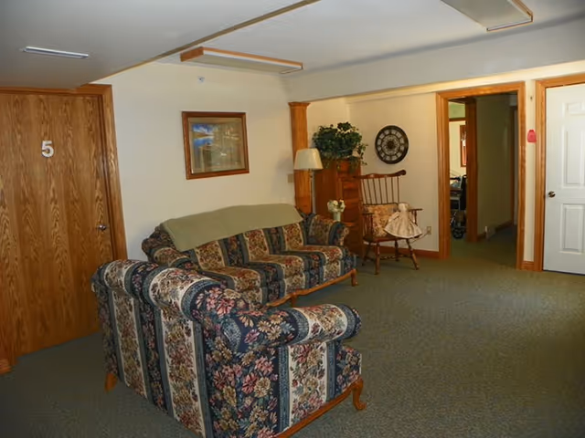 A cozy living room area with floral patterned sofas, a wooden rocking chair with a doll seated on it, a wooden door labeled with the number 5, a framed picture on the wall, a floor lamp, and a decorative clock. The room has beige walls and carpeted flooring.