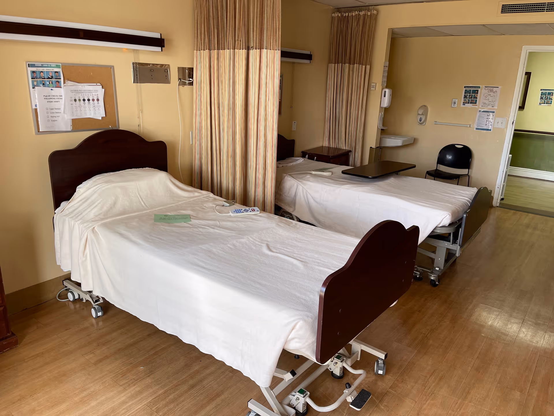 A room with two hospital-style beds separated by a privacy curtain. Each bed has white sheets and wooden headboards and footboards. There is a small table on wheels next to one bed, a chair against the wall, a sink with soap dispensers, and various notices and charts pinned on the walls. The floor is wooden, and the room has a warm beige color.