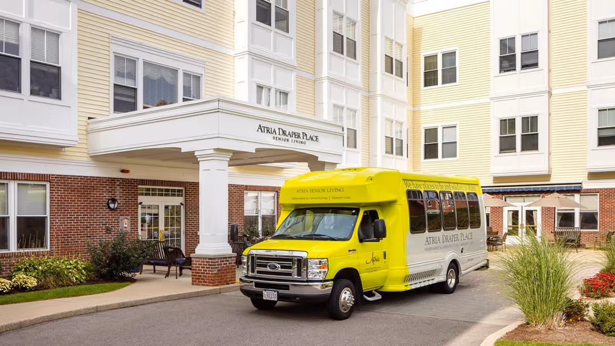 Exterior view of Atria Draper Place senior living facility showing the building entrance with a covered drop-off area and a yellow shuttle bus parked in front. The building has multiple windows and a combination of brick and light yellow siding. There are some plants and outdoor seating visible near the entrance.
