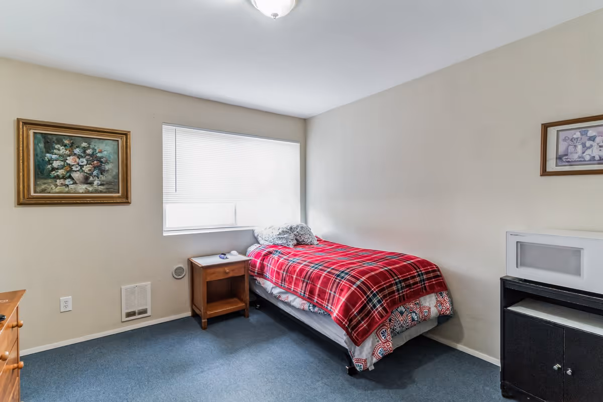 Small single bedroom with a twin bed covered by a red plaid blanket, a wooden nightstand under a window with blinds, framed wall art, and a cabinet with a microwave.