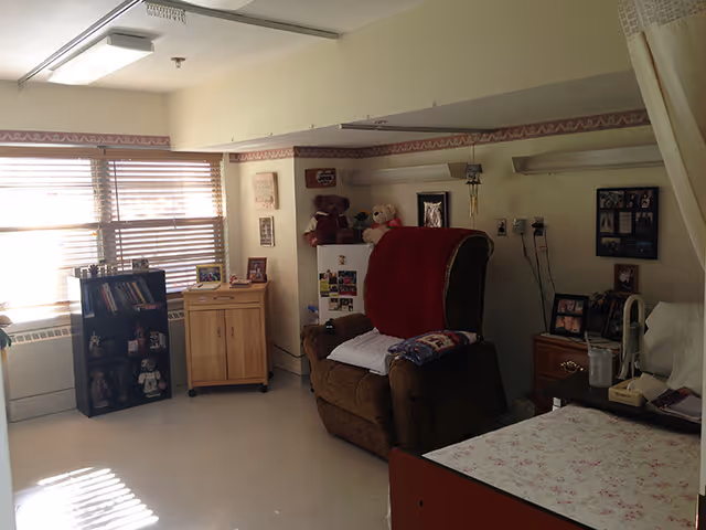 A cozy room with a large brown recliner chair covered with a red blanket and a white pillow. There is a small wooden cabinet and a black bookshelf filled with books and decorative items near a window with blinds. The room also contains a hospital-style bed with a floral bedspread, a nightstand with framed photos, a glass of water, and a telephone. The walls have some framed pictures and teddy bears placed on a shelf.