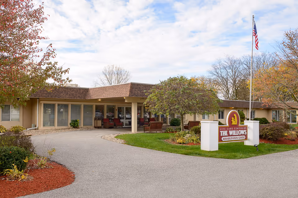 Exterior view of Life Care Center of The Willows Rehabilitation Center showing a single-story building with a covered entrance, outdoor seating, a flagpole with an American flag, and landscaped greenery including trees and shrubs.