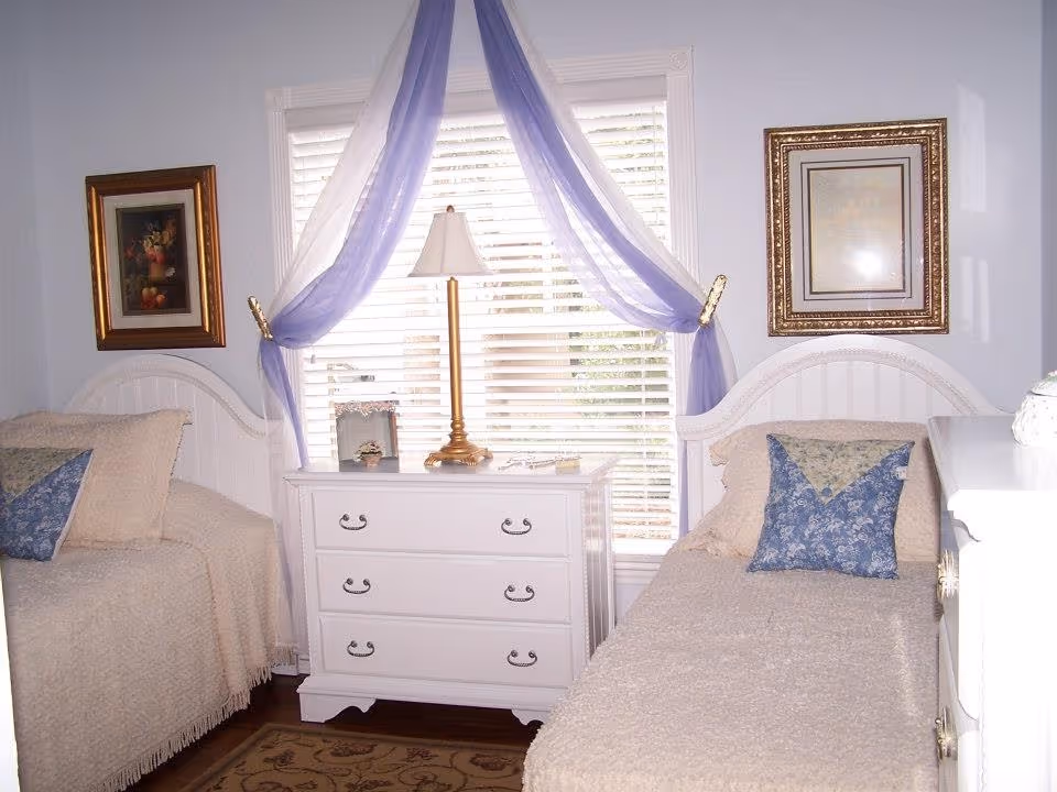 A bedroom with two twin beds separated by a white three-drawer dresser. Each bed has a beige blanket and a decorative blue pillow. A table lamp with a gold base sits on the dresser in front of a window with white blinds and sheer purple curtains draped on either side. Two framed pictures hang on the light blue walls above each bed.