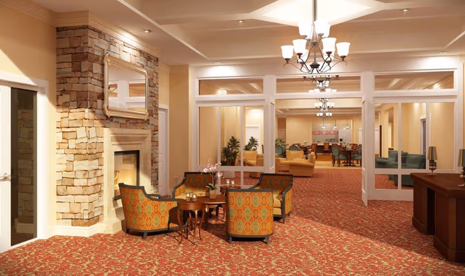 A spacious and warmly lit senior living facility common area with patterned red carpet, a stone fireplace with a mirror above it, and a seating arrangement of four upholstered chairs around a small wooden table. The room extends into another area with more seating and tables, illuminated by multiple chandeliers and lamps.