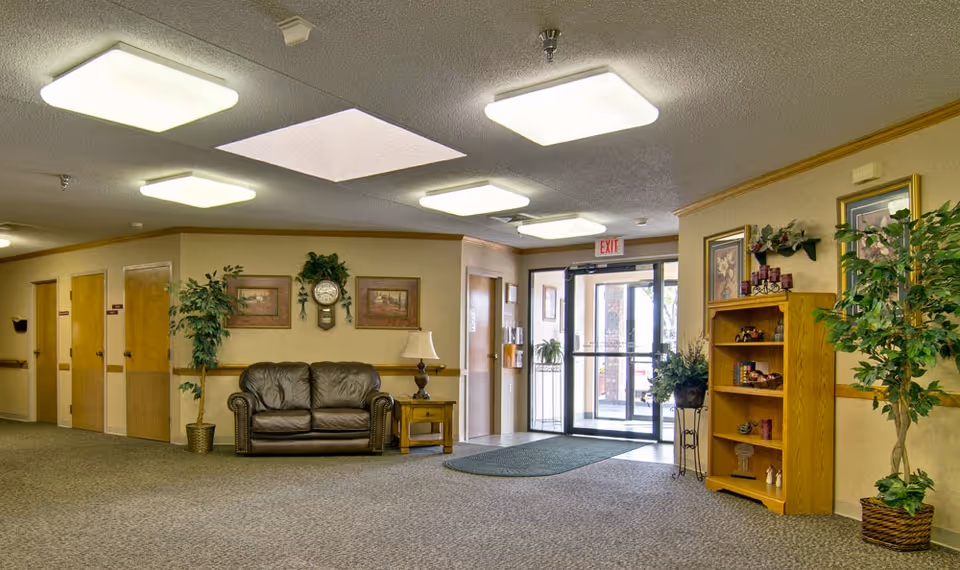 Interior view of a nursing center lobby area with a brown leather loveseat, wooden side table with a lamp, potted plants, framed artwork, a clock on the wall, a wooden bookshelf with decorative items, and glass double doors leading outside with an exit sign above.