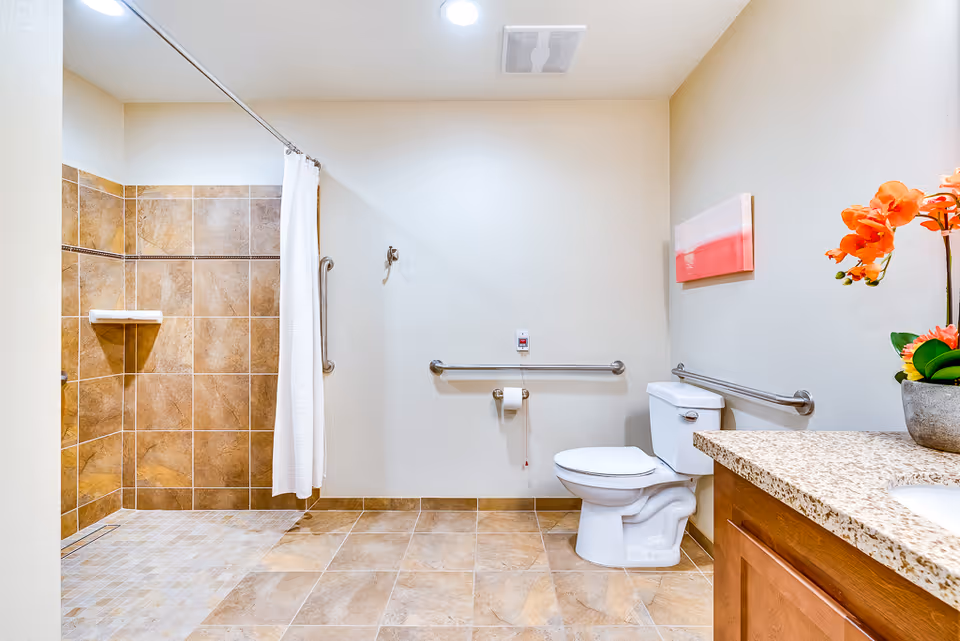 A spacious senior living bathroom with beige tiled floors and walls, featuring a walk-in shower with a white curtain, a white toilet with grab bars on the walls, a granite countertop with a sink, and a potted plant with orange flowers on the counter. There is also a small piece of wall art above the toilet.