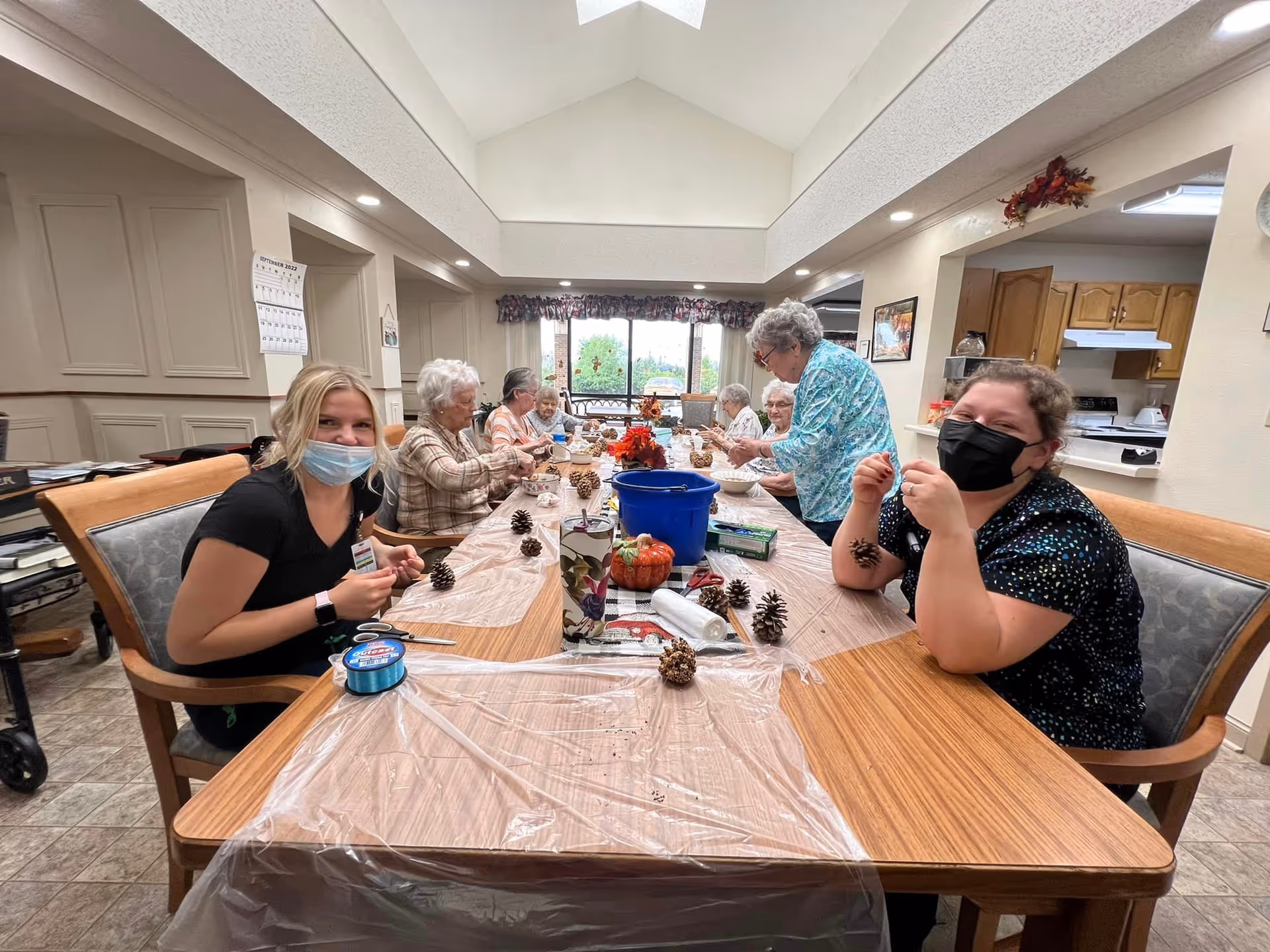 A group of elderly women and two caregivers wearing masks are seated around a long table in a well-lit room, engaging in a craft activity involving pinecones. The table is covered with a plastic sheet and decorated with small pumpkins and fall-themed items. The room has a high ceiling with skylights and an open kitchen area in the background.