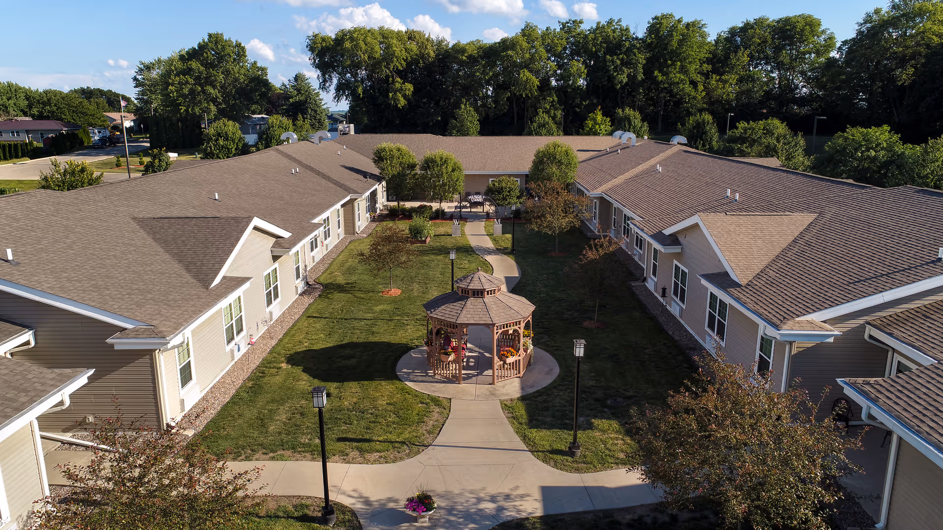 Aerial view of a senior living facility courtyard with a central gazebo surrounded by a well-maintained lawn, trees, and pathways. The courtyard is enclosed by single-story buildings with beige siding and brown roofs. The sky is clear with a few clouds, and there are lamp posts along the pathways.