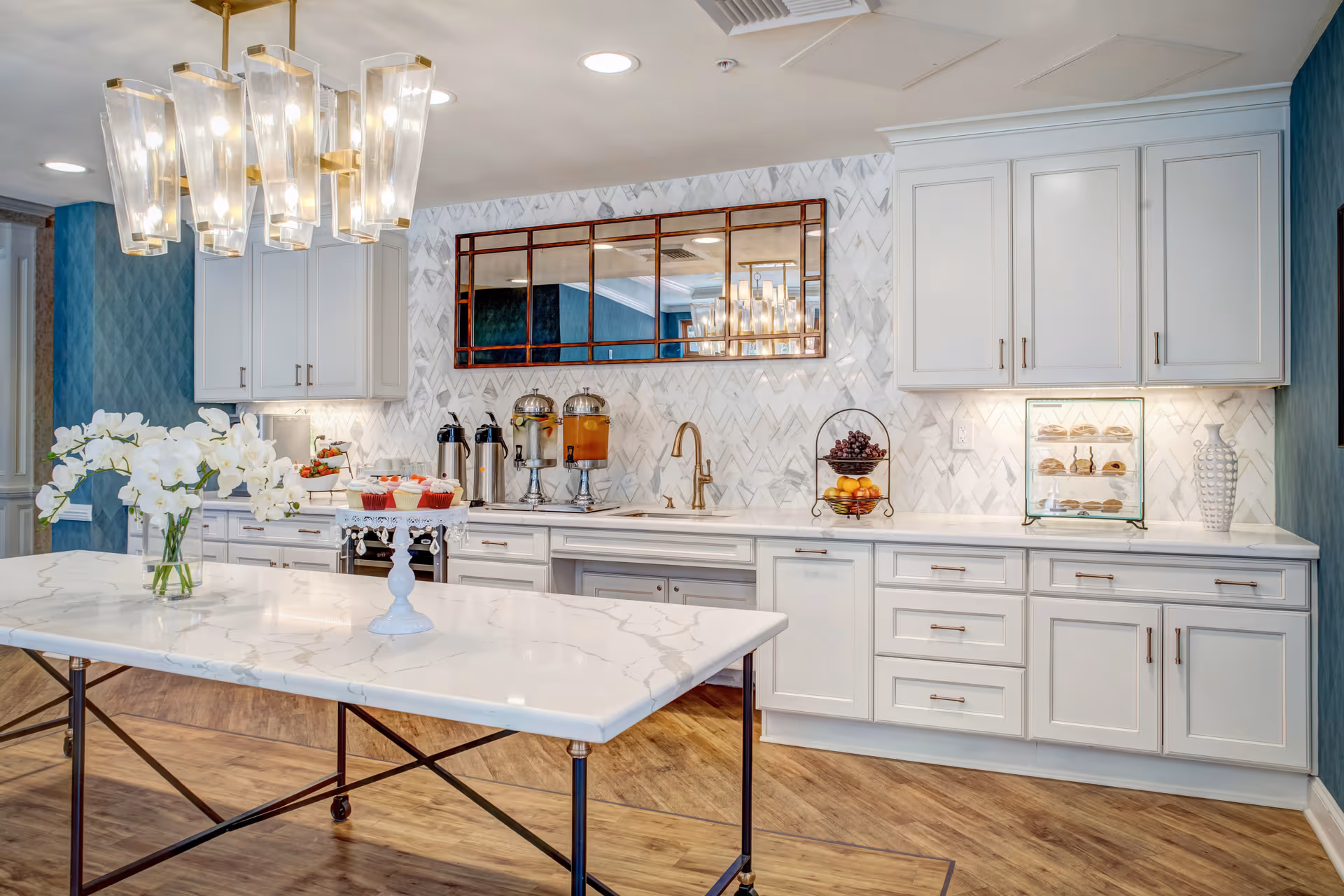 Bright modern kitchen/buffet area with a marble island, white cabinets, beverage dispensers and pastries under warm pendant lighting.