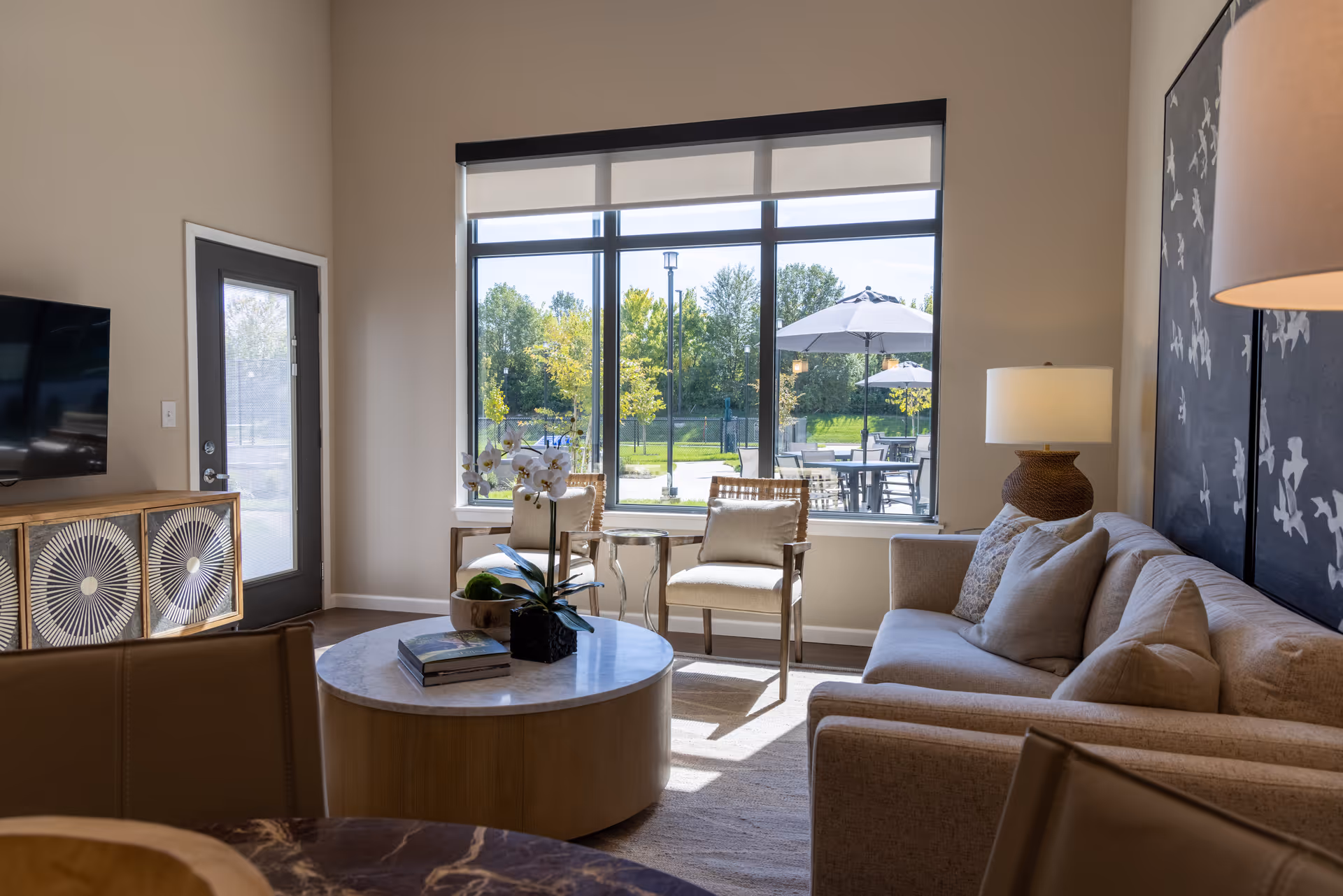 A bright and modern living room with a beige sofa, two wooden chairs with cushions, a round wooden coffee table with books and a plant, a large window showing an outdoor patio with tables and umbrellas, a TV on a decorative wooden cabinet, and a floor lamp next to the sofa.
