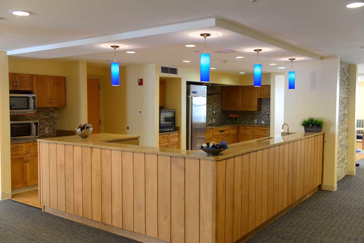 Interior view of a kitchen area in a senior living facility featuring wooden cabinets, a stainless steel refrigerator, a built-in microwave, and a countertop with a wooden panel front. The ceiling has recessed lighting and four blue pendant lights hanging above the counter. There are decorative bowls on the counter and a potted plant on the right side. The floor is carpeted outside the kitchen area.
