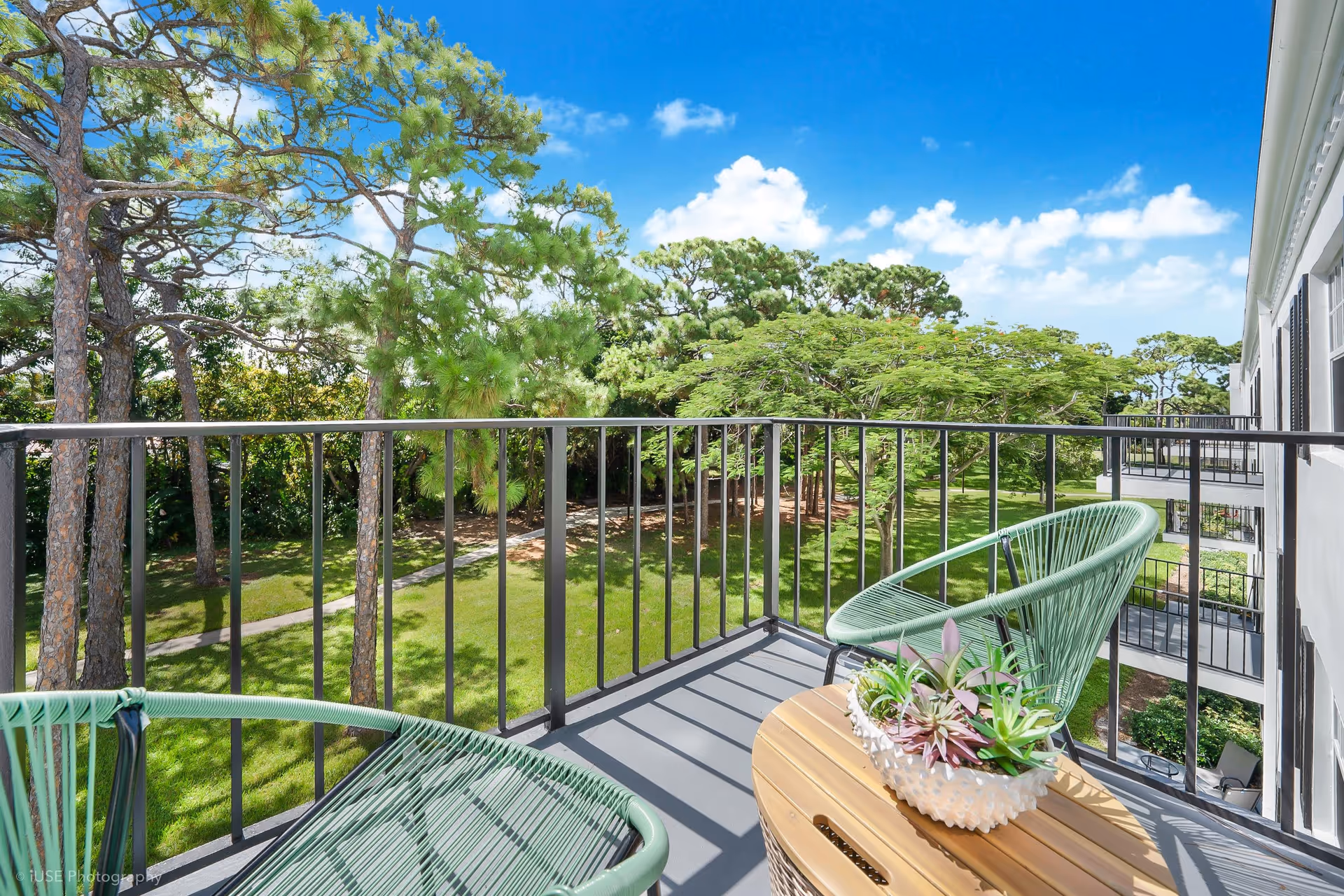 Balcony with two green chairs and a small table overlooking a grassy courtyard and trees under a blue sky.