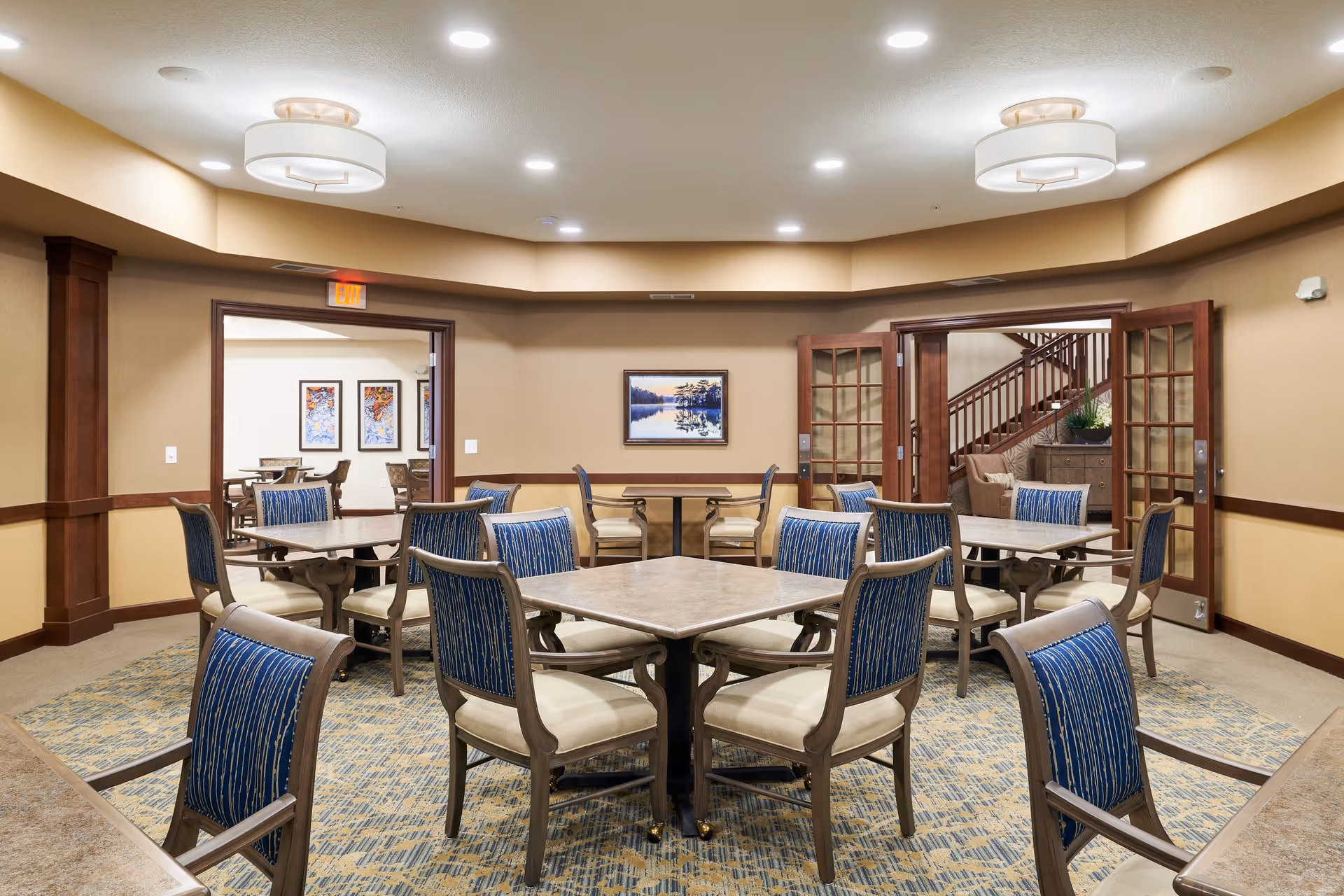 A bright communal dining room with several square tables and blue-upholstered chairs in a senior living facility.