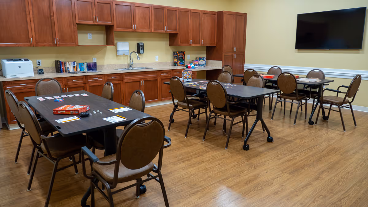 A community room with three tables and several chairs arranged around them. On the tables are board games and game pieces. The room has wooden cabinets along one wall with a sink, a printer, and books. A large flat-screen TV is mounted on the opposite wall. The floor is wood, and the walls are painted a light yellow.