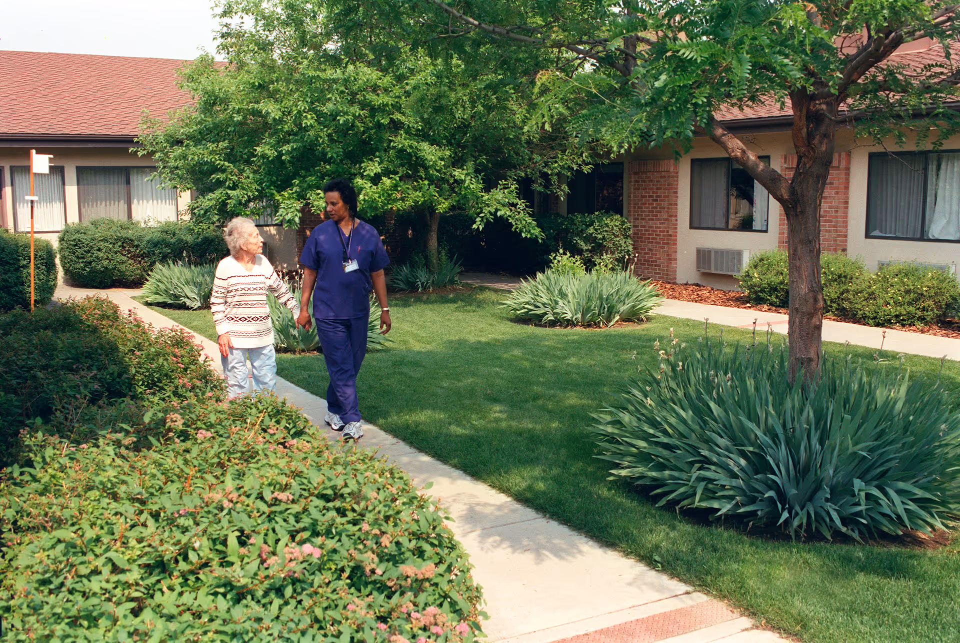 An elderly woman and a caregiver walking together on a sidewalk in a garden area of a senior living facility. The garden is lush with green grass, bushes, and trees, and the building with windows and a red roof is visible in the background.