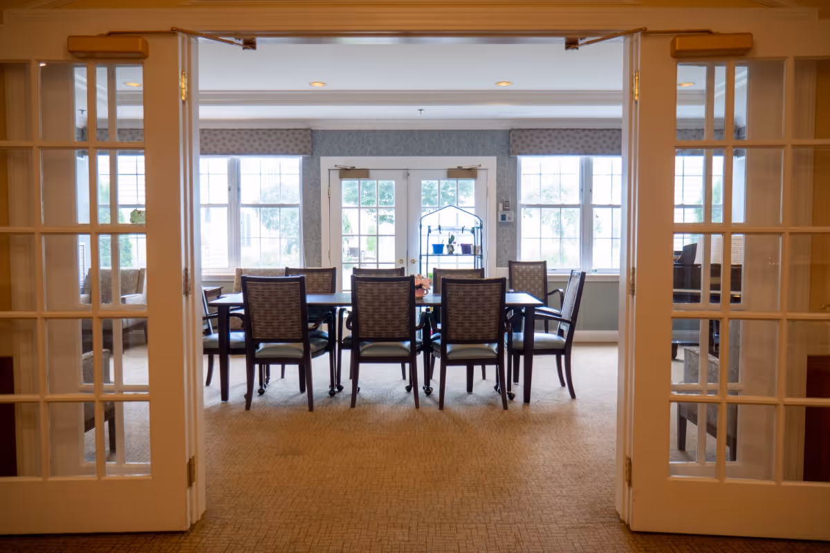 Dining room with a long table and several chairs viewed through open French doors and large windows.