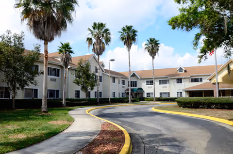 Exterior view of a senior living facility with a curved driveway, palm trees, and a two-story building with beige siding and a brown roof under a partly cloudy sky.