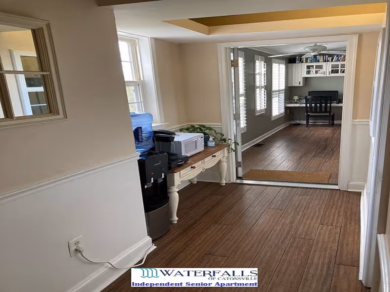 Hallway interior showing a console table with a water cooler, microwave and coffee maker leading into a sunlit room with a desk and chair.