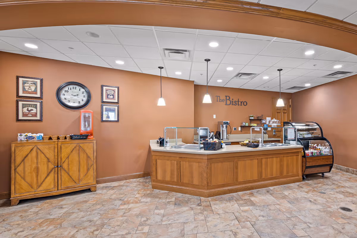 Interior view of The Bistro at The Deerfield, featuring a wooden service counter with glass sneeze guards, a refrigerated display case with beverages and snacks, and a wooden cabinet against a brown wall with a clock and framed coffee-themed artwork. The floor is tiled, and pendant lights hang from the ceiling.