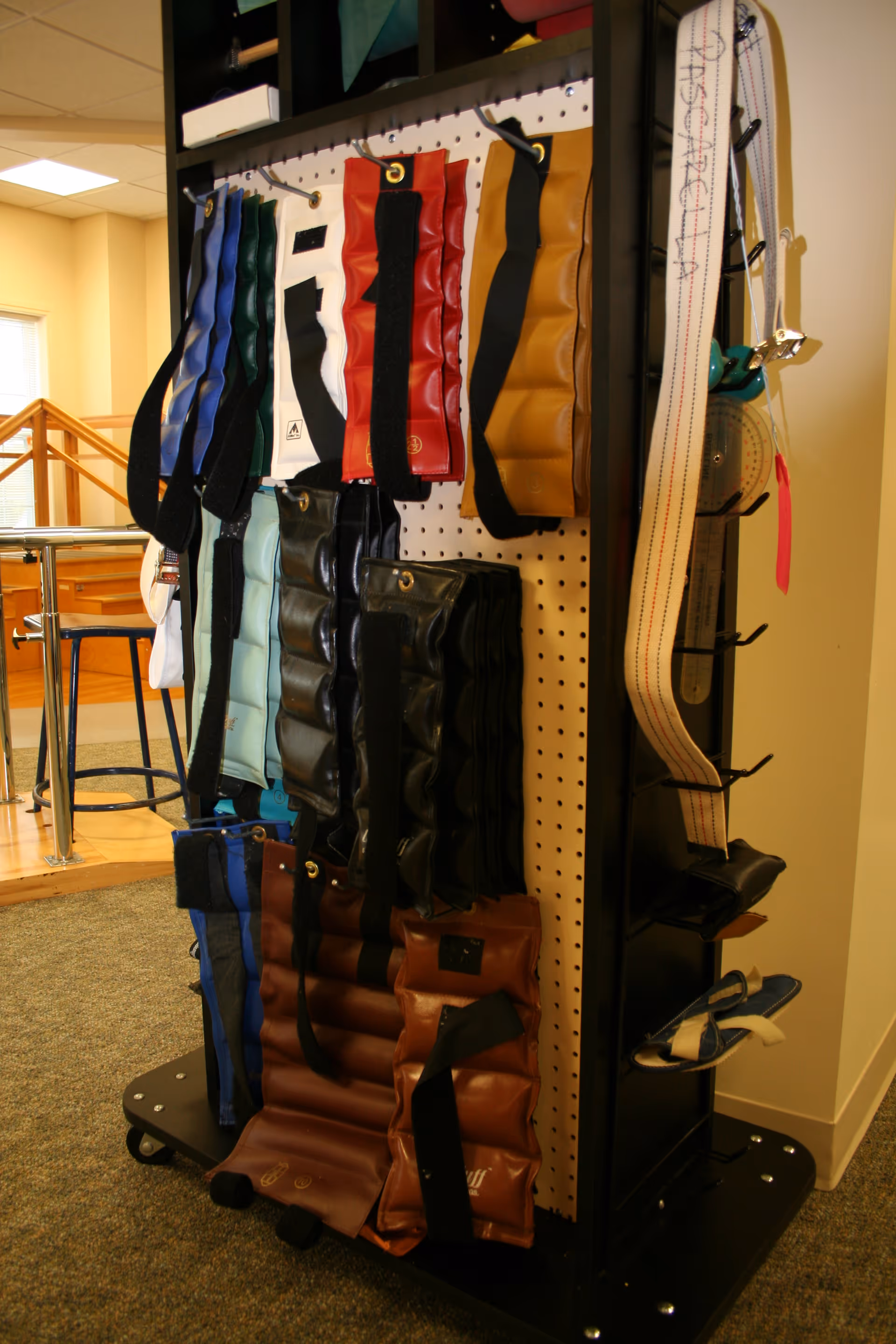A black pegboard stand displaying various colored weighted ankle or wrist cuffs used for physical therapy or rehabilitation, located in a room with carpeted floor and wooden stairs in the background.