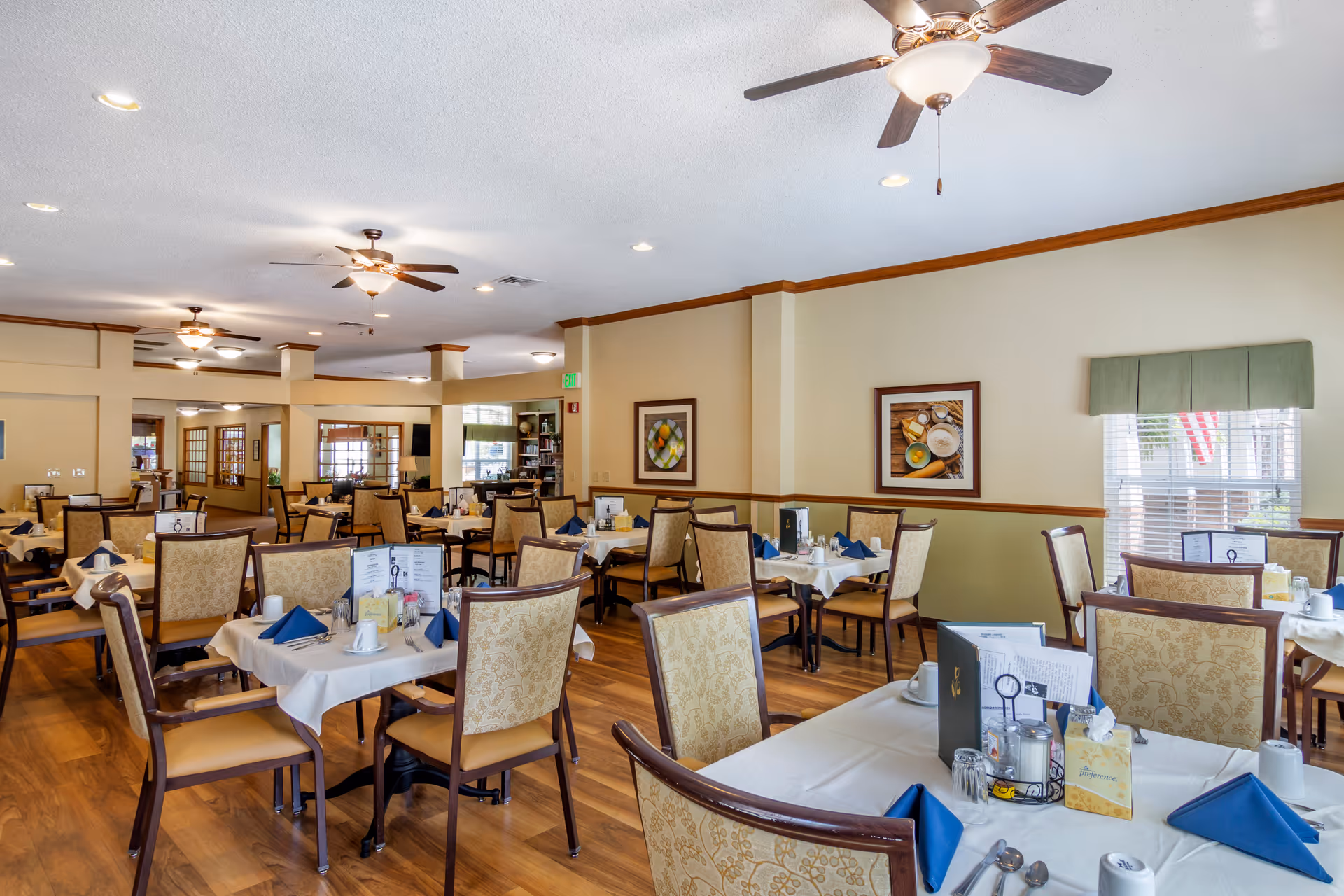 Spacious dining room with multiple set tables and upholstered chairs, ceiling fans, and framed wall art.