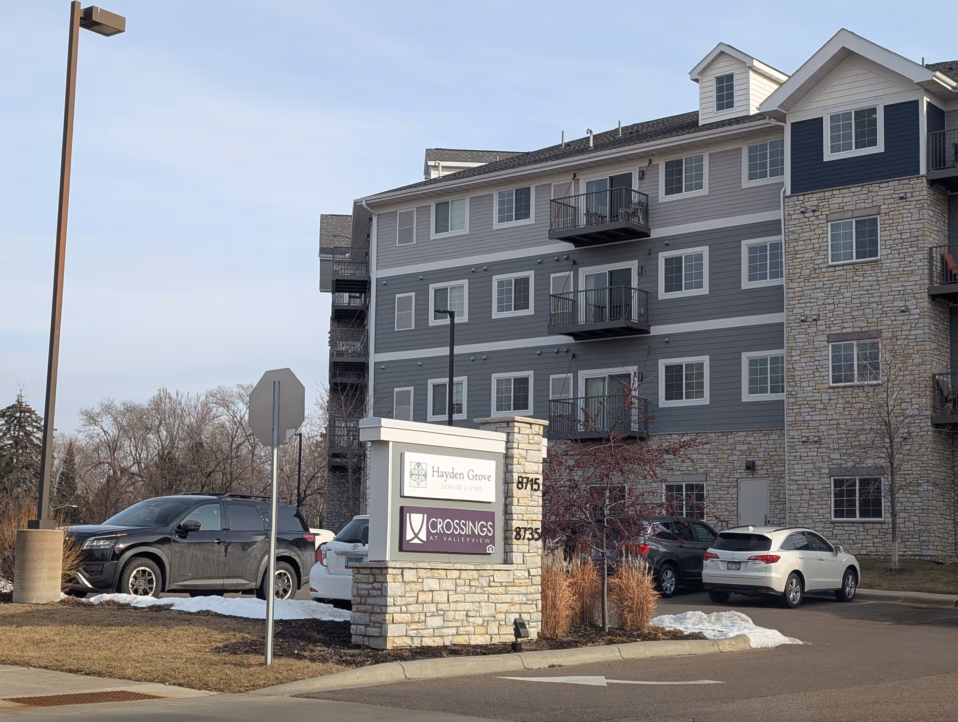 Exterior view of a multi-story senior living facility building with stone and siding facade. There is a parking lot with several cars and a sign in front that reads 'Hayden Grove Senior Living' and 'Crossings at Valleyview'. The sky is clear and there are some leafless trees around.