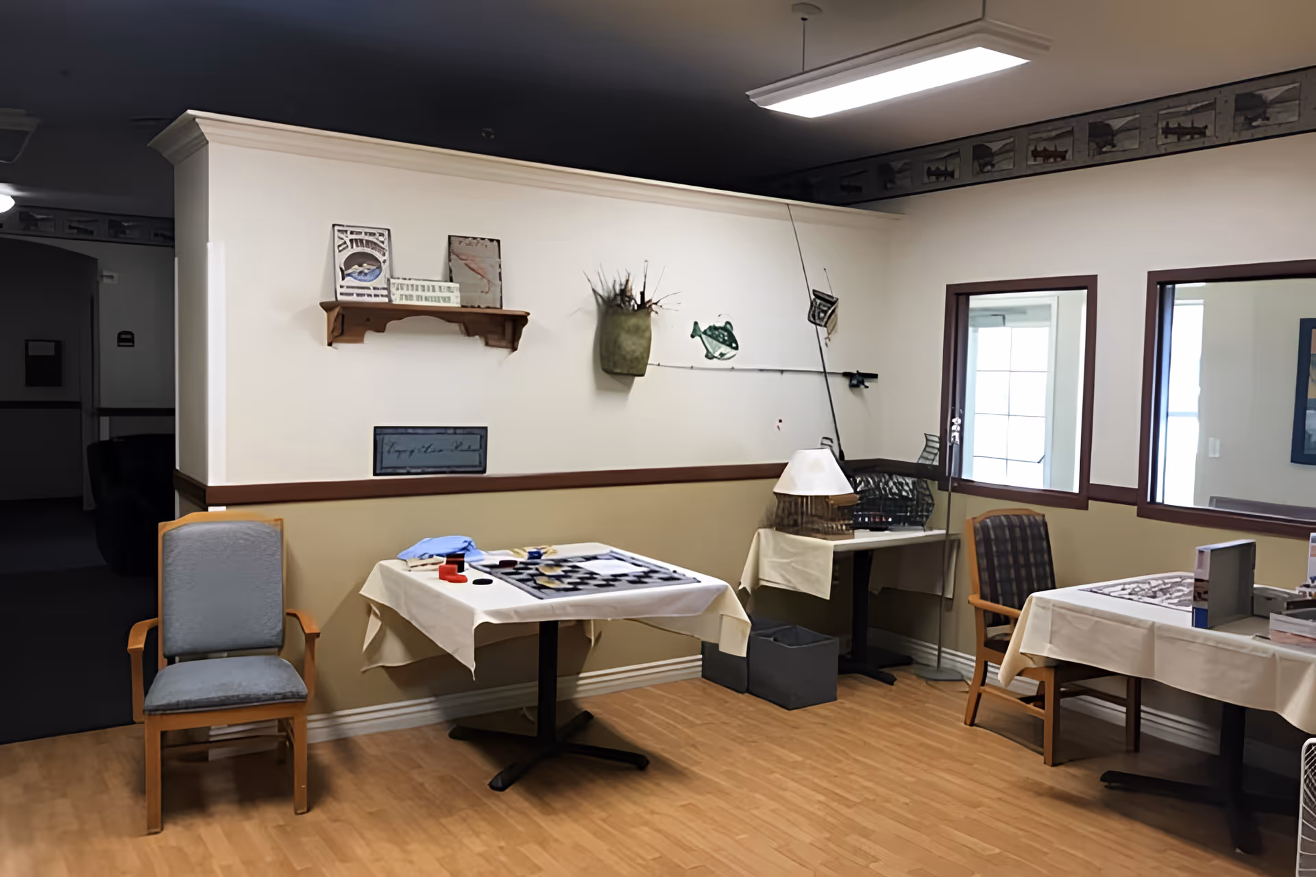 A cozy interior room with wooden flooring and beige walls, featuring two tables covered with white tablecloths. One table has a checkerboard game set up, and the other has various items on it. There are two chairs, one with a blue cushion and the other with a plaid cushion. The walls are decorated with a small wooden shelf holding framed pictures and a wall-mounted vase with decorative branches. A fishing rod and fish-themed decorations are also mounted on the wall. Two windows with dark brown frames look into another room.