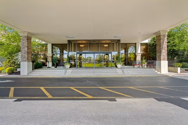 Entrance of a building with a covered drop-off area, glass doors, stone pillars, and outdoor seating on either side. Green trees and landscaping are visible around the entrance.